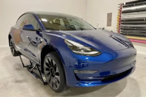 Blue Tesla Model 3 with black wheels and tinted windows, showing the front view lifted on a jack.
