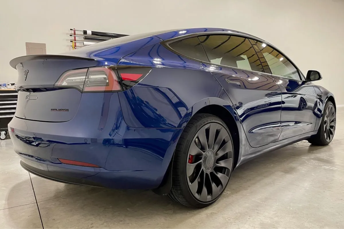Blue Tesla Model 3 with black wheels and tinted windows, showing the rear view with a clear reflection.